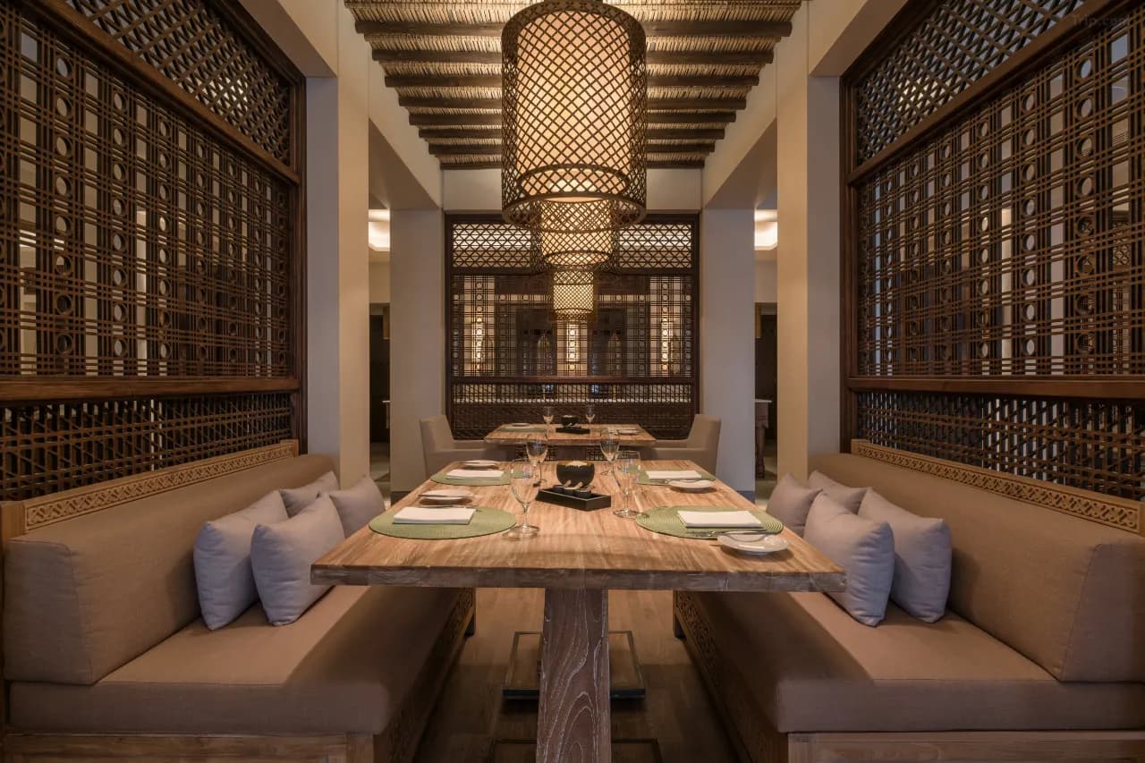 An elegant dining room with a long wooden table set for a meal, flanked by built-in bench seating and decorative dark wood mashrabiya lattice screens under large woven pendant lights.