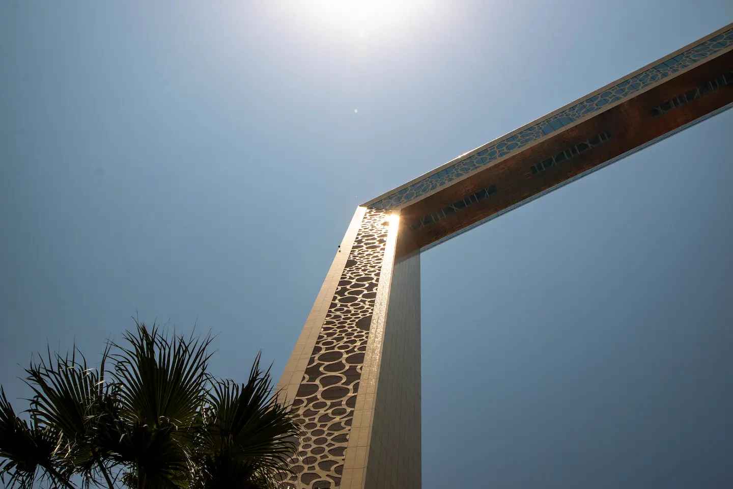 A low-angle perspective of one corner of the Dubai Frame, capturing the sun's reflection on the golden cladding and the contrast between the structure and the deep blue sky.