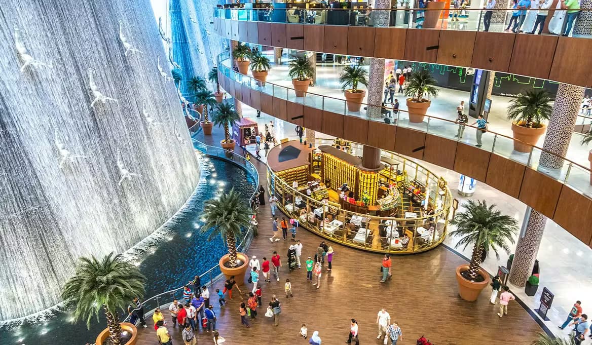 The Dubai Mall indoor waterfall atrium with visitors, palm trees, and a restaurant on multiple floors