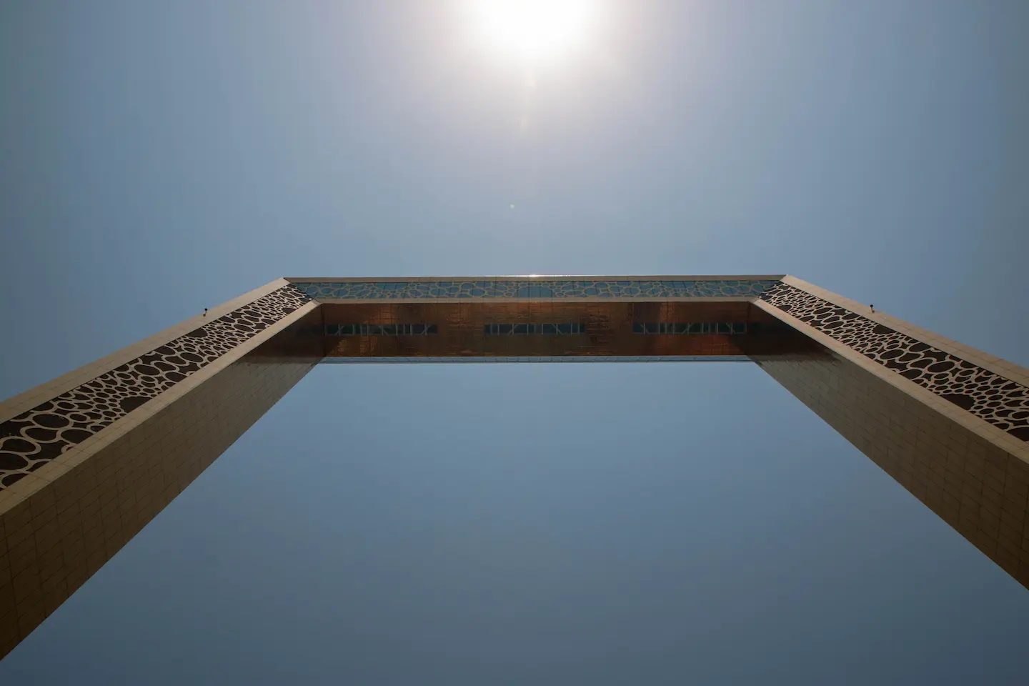 A dramatic low-angle shot looking directly up at the Dubai Frame, highlighting the intricate circular patterns on the golden pillars and the sun shining brightly above the sky bridge.