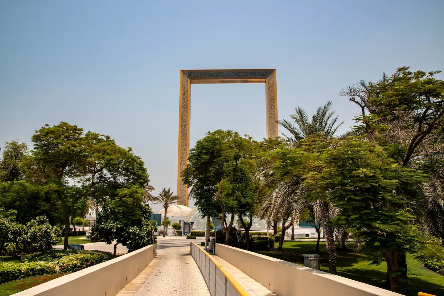 A paved walkway in Zabeel Park lined with palm trees and greenery, leading toward the towering Dubai Frame landmark under a bright midday sun.