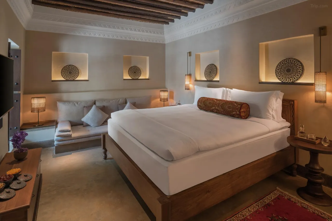 A cozy, heritage-inspired bedroom with a solid wood bed frame, white linens, and a patterned bolster pillow. The walls feature recessed niches with decorative metal plates and warm backlighting.
