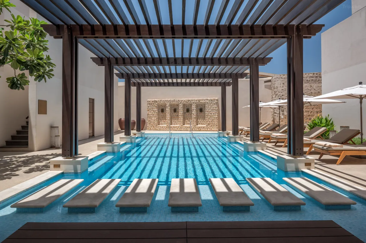 A serene outdoor swimming pool covered by a large dark wood pergola, creating linear shadows on the water. The area includes submerged lounge ledges and white umbrellas in the background.