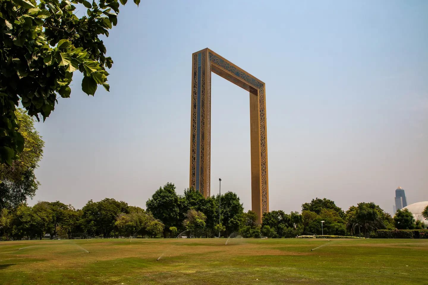 The Dubai Frame seen from a lush green lawn in Zabeel Park, showing its gold-patterned exterior and the observation bridge at the top against a clear sky.